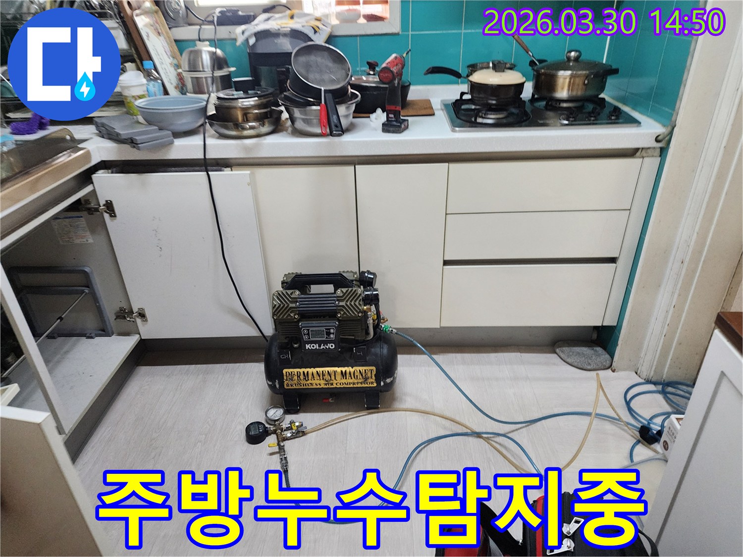 주방 하부장 문이 열린 채-누수 탐지용 공압 컴프레서가-배관에 연결되어 작동하고 있습니다.