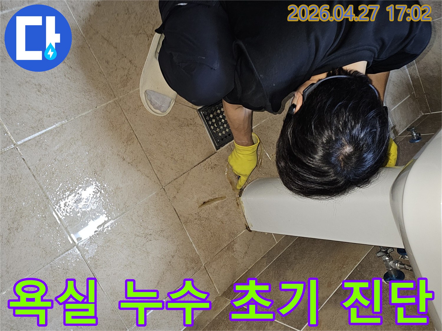 누수 전문가가-노란 장갑을 끼고-욕실 바닥의 젖은 타일과-세면대 하단부를-집중적으로 관찰하며-누수 지점을 파악하고 있습니다