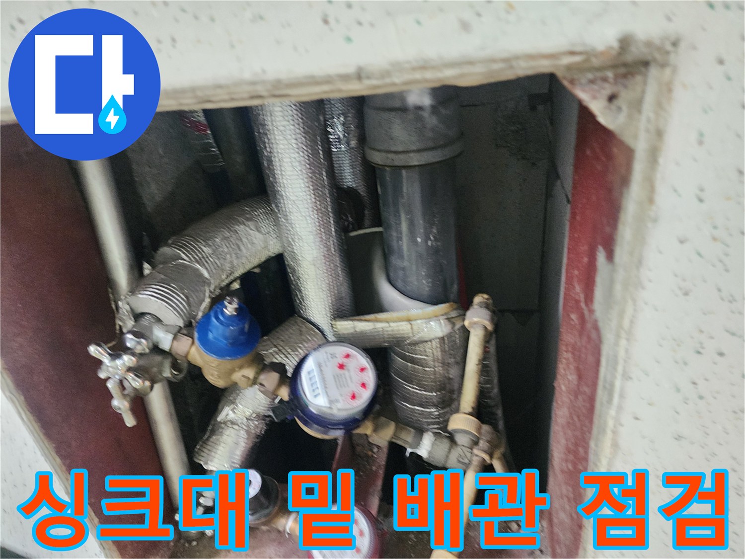 보온재가 씌워진 배관과 수치 표시 계량기가 설치된 주방 하단 내부 모습입니다.
