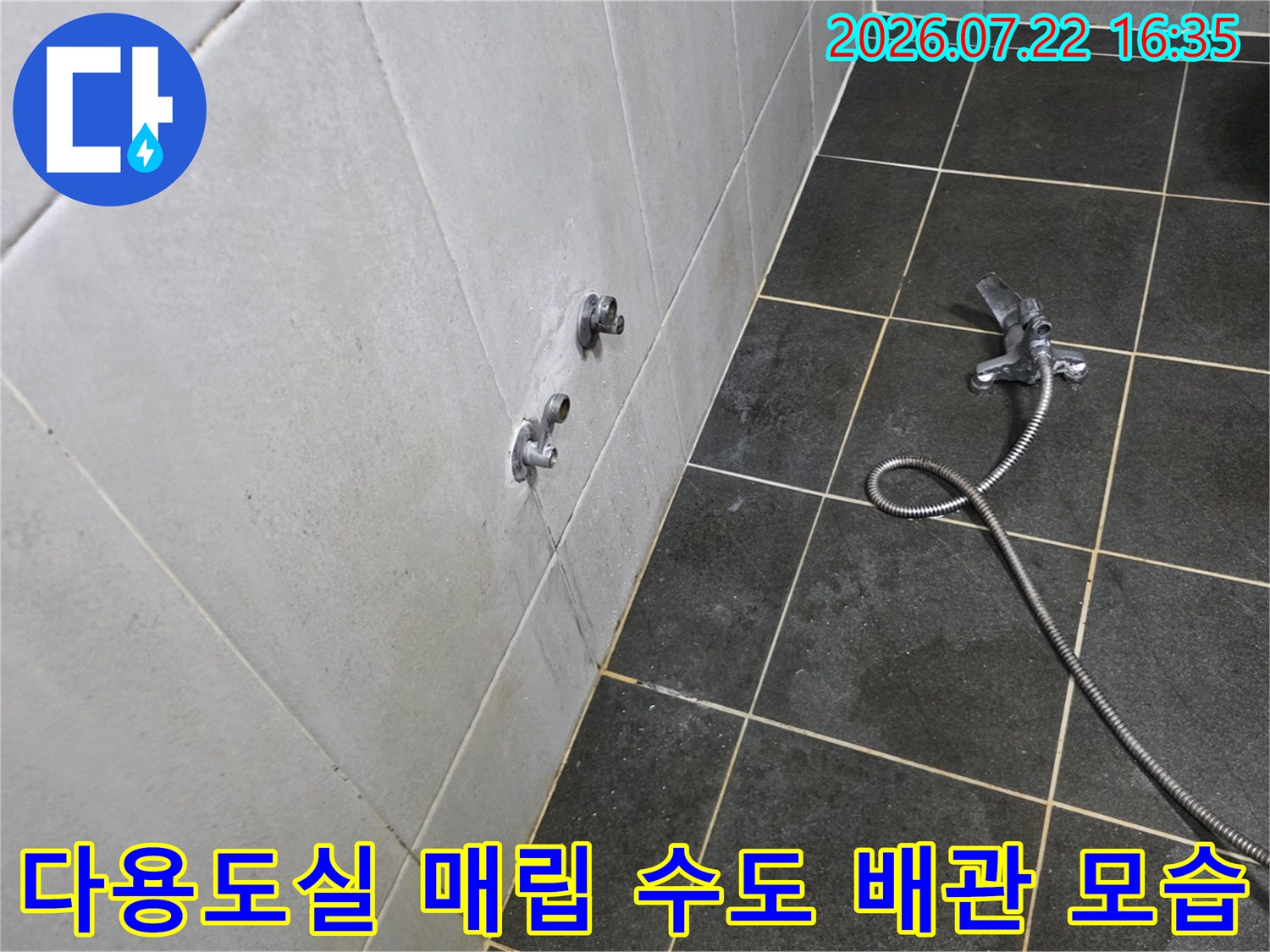 어두운 회색 바닥 타일 위에 샤워 호스가 놓여 있고 벽면에 수전이 돌출된 세탁실 전경